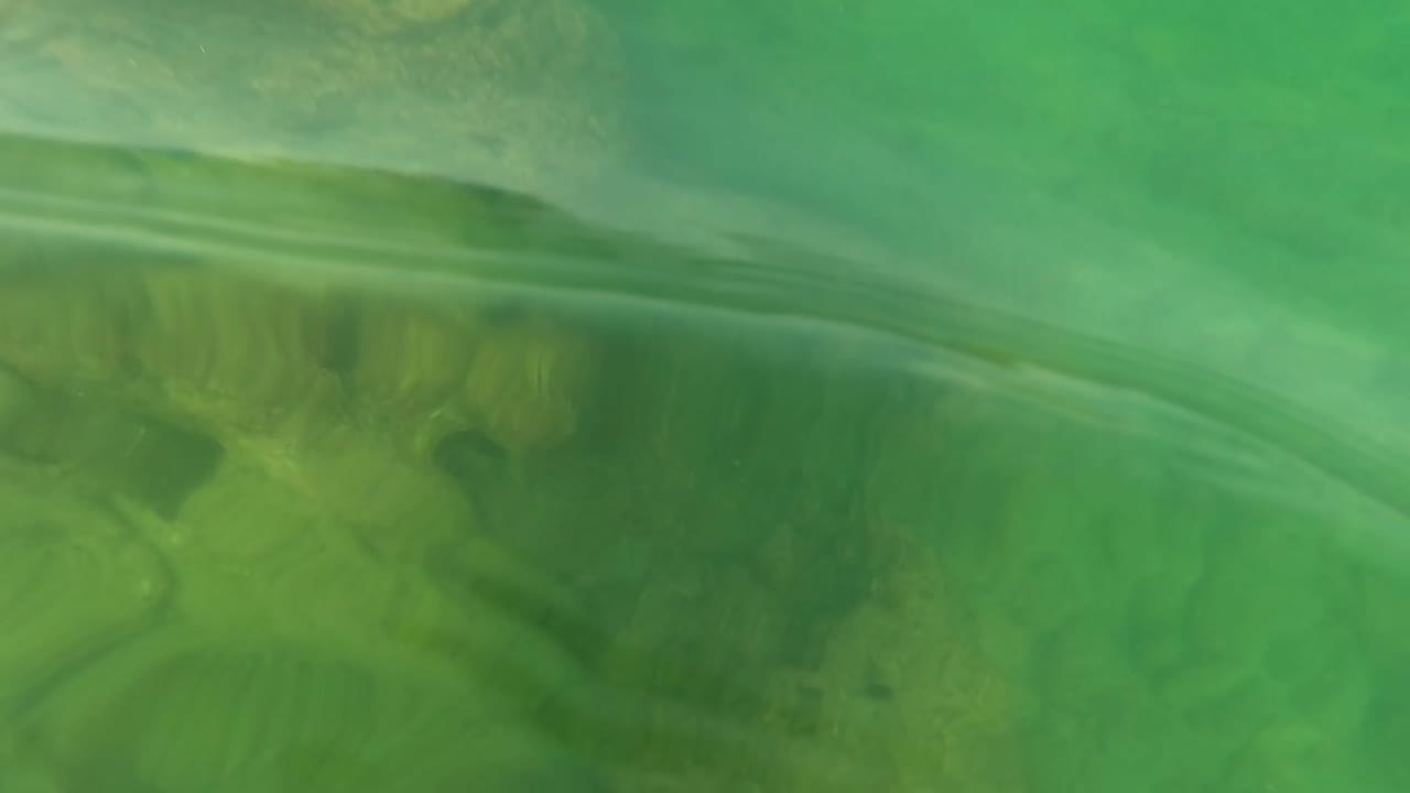 Waterline view in Los Barriles showing green water, a curved surface ripple, and darker submerged rock forms below, no visible people.