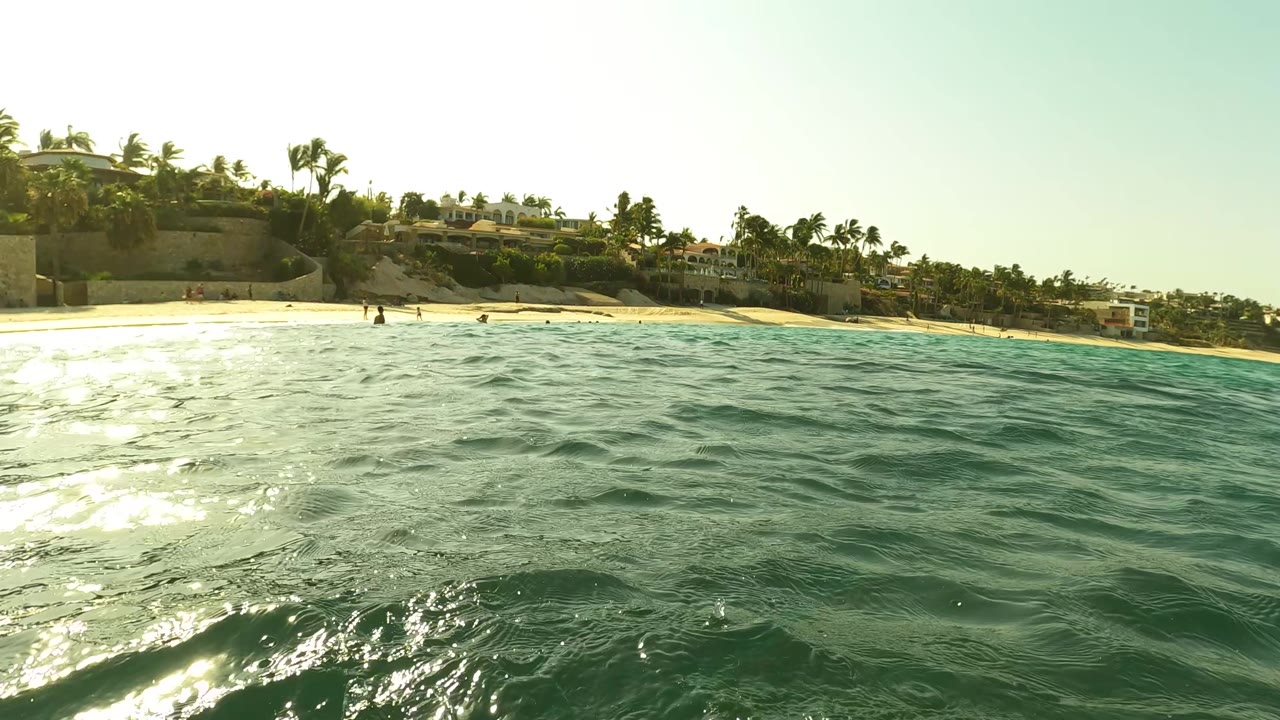 Waterline view in Los Barriles, Baja California Sur, looking across rippled ocean toward a sandy beach with palm trees, buildings, and tiny distant people.