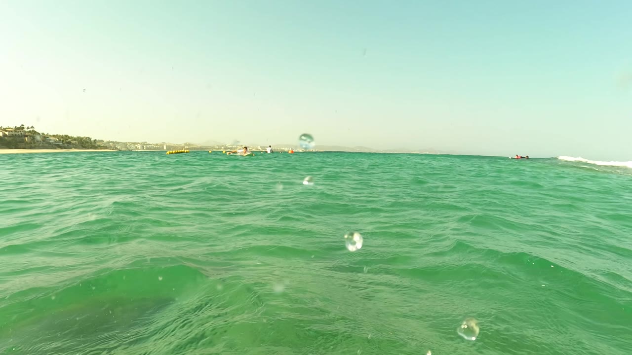 Waterline view in Los Barriles, Baja California Sur, Mexico, with rippled blue-green ocean in the foreground, distant shore and boats on the horizon, and small people visible near the shore.