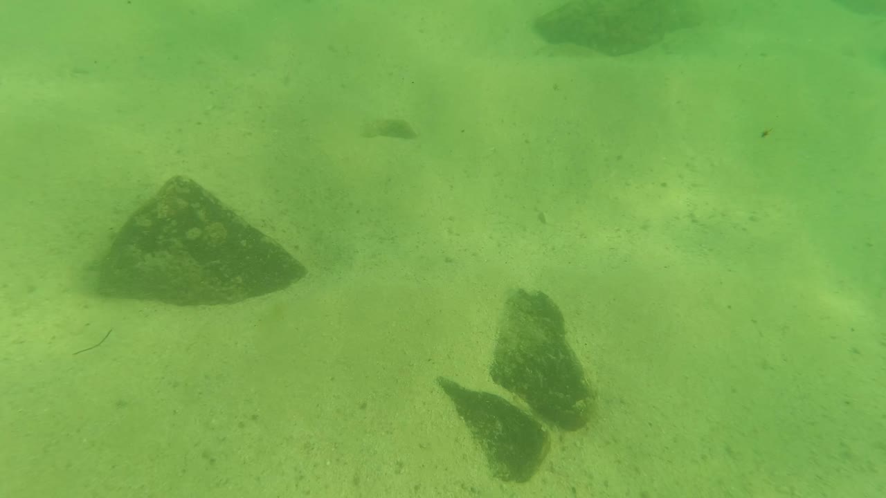 Underwater view in Los Barriles, Baja California Sur, with larger rocks on the sandy bottom, a soft green cast through the water, and no visible people.