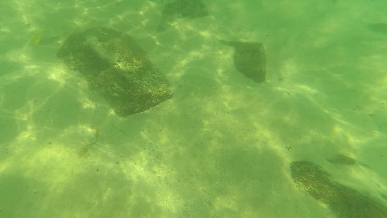 At 0:02 in Los Barriles, Baja California Sur, dark rocks sit on a pale sandy bottom beneath clear green water with bright ripple patterns, with no visible people.