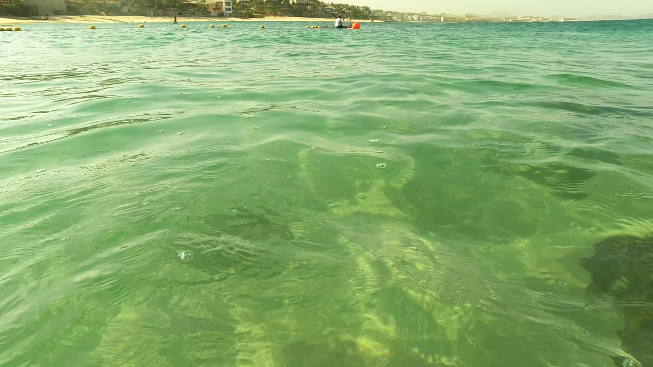 Water-level view of shallow green ocean near Los Barriles, Baja California Sur, with rippled surface, visible seabed shapes, distant swimmers, buoys, and shoreline along the top of frame.