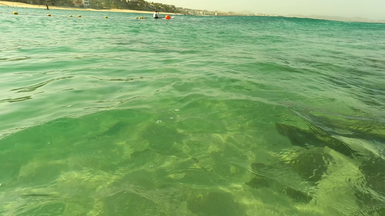 Shallow green ocean near Los Barriles, Baja California Sur, shows broad ripples and underwater rock or seabed forms, with distant buoys, swimmers, and shoreline compressed along the upper edge.