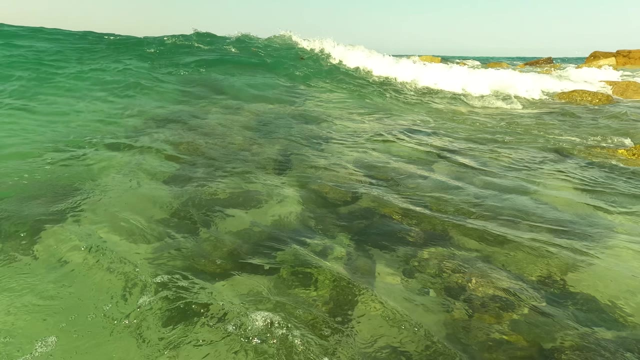 At Los Barriles, Baja California Sur, the camera remains at water level as a green swell rises across the frame and white foam breaks over rocks, with no visible people.