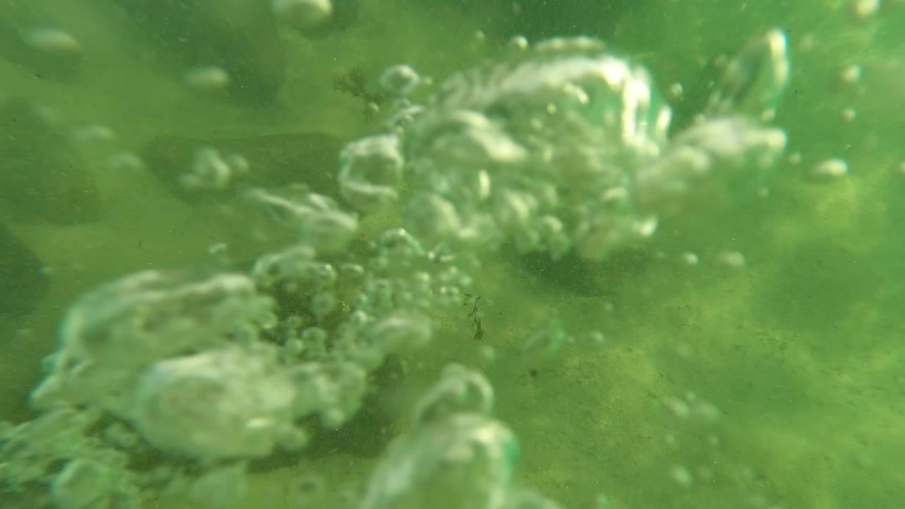 At Los Barriles, Baja California Sur, the view is underwater with large bubbles in the foreground and muted rocks and sandy bottom behind them, with no visible people.