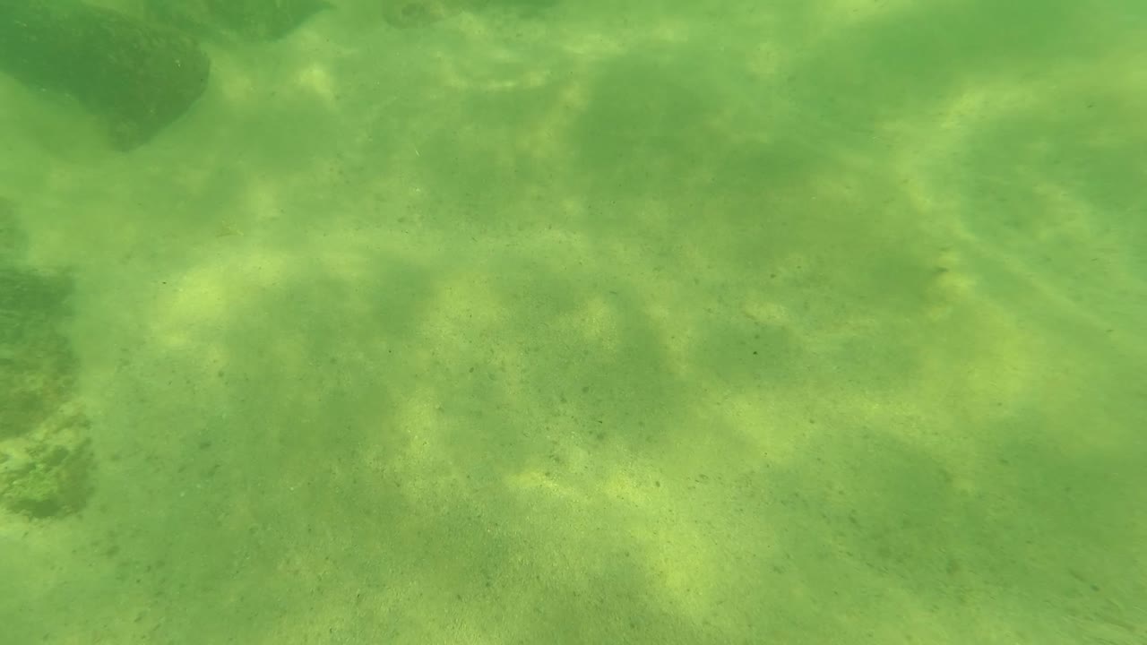 Mid-clip underwater frame from Los Barriles, Baja California Sur showing a pale sandy bottom, dark rocks near the top and lower edge, green water, and no visible people.