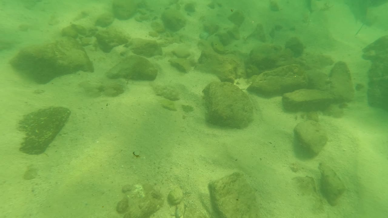 Second underwater frame in Los Barriles, Baja California Sur, with larger rocks near the upper edge, pale sand below, and no visible people.