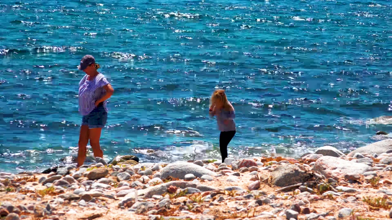 Adult in a striped shirt and child in a light top walk apart on the rocky shoreline in La Ventana, Mexico, with bright blue water behind them.