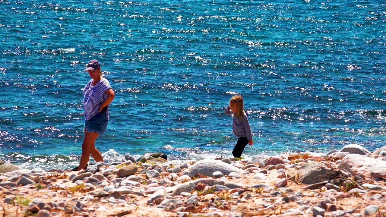 Adult walking near a child on rocks with waves behind them.