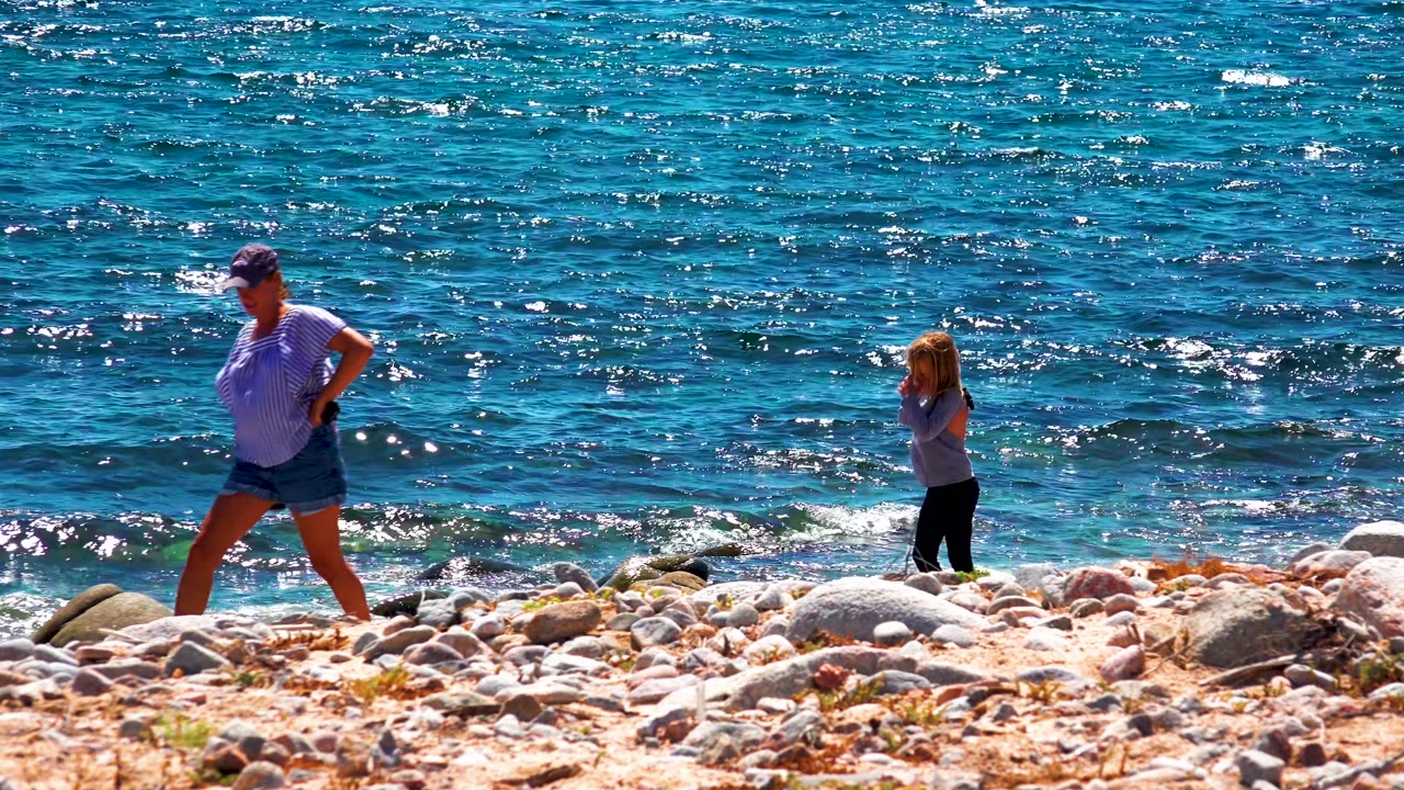 The adult faces across the rocky beach while the child appears nearer the breaking waves, with sparkling blue water covering the background in La Ventana, Mexico.