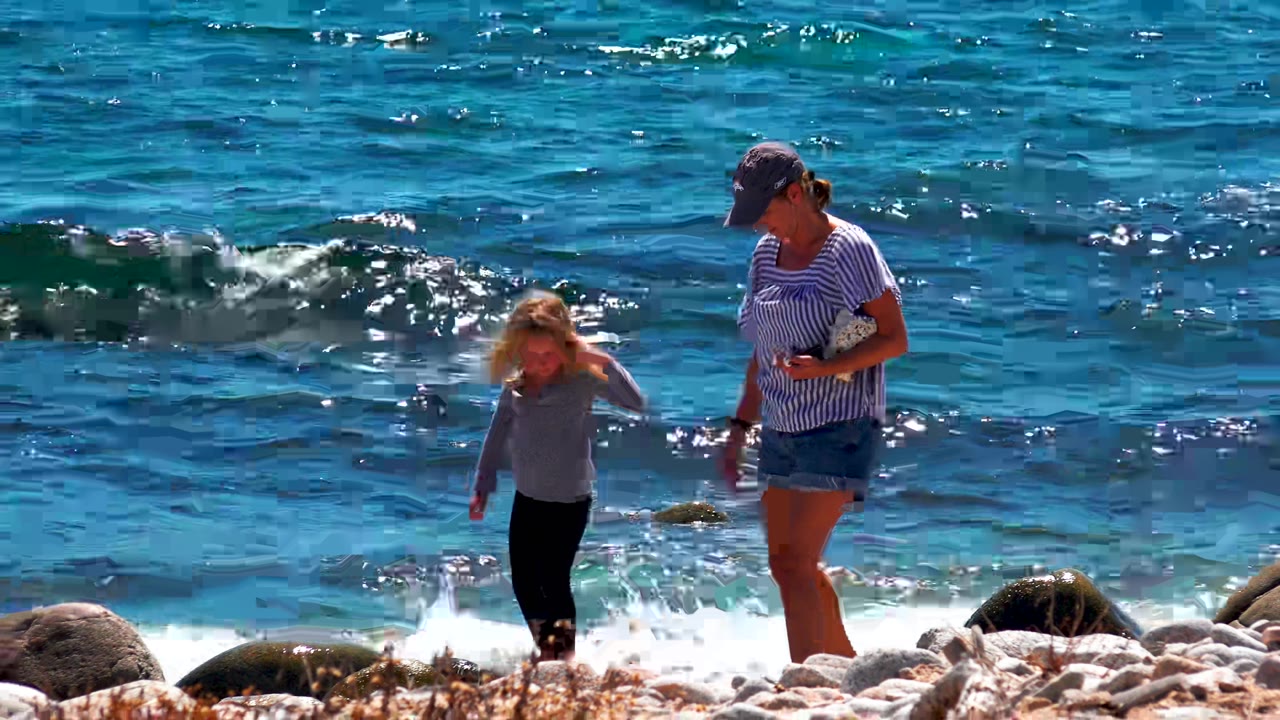 At 0:00 in La Ventana, Mexico, an adult in a striped shirt and cap walks beside a child near rounded shoreline rocks as blue waves break behind them.