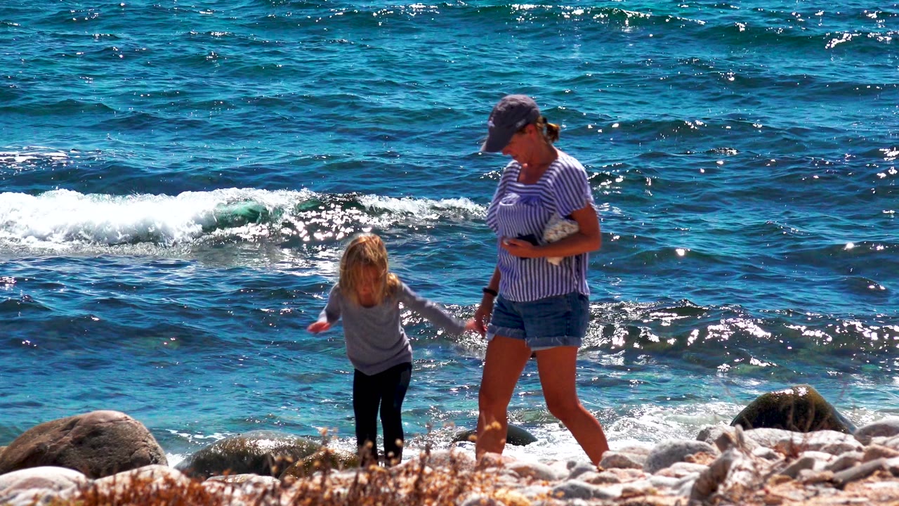 Child and adult wading near rocks with a white wave breaking behind them.