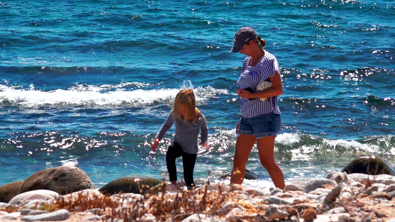 At 0:02 in La Ventana, Mexico, the adult looks down while standing beside the child, and a low white wave curls across the midground behind shoreline rocks.