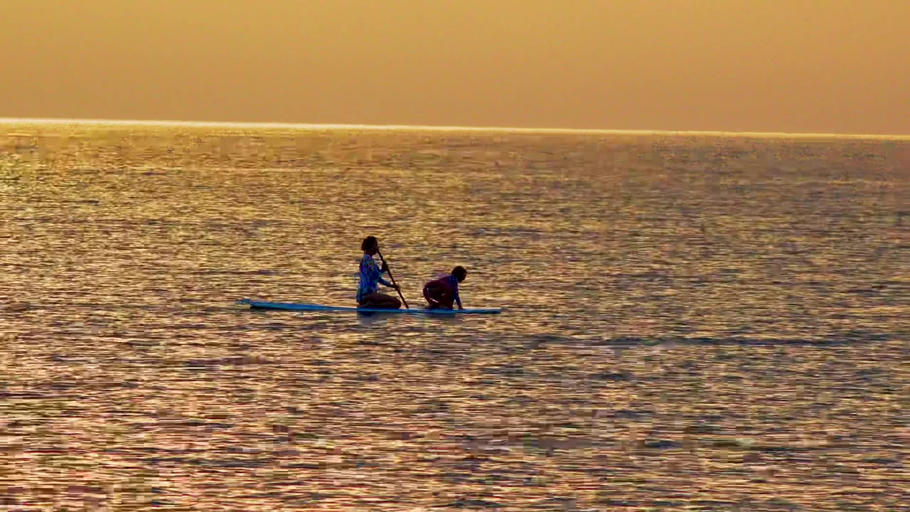 Two people share a paddleboard on the ocean at sunset in El Sargento La Ventana, Mexico, with one kneeling near the center and the other bent low near the front.