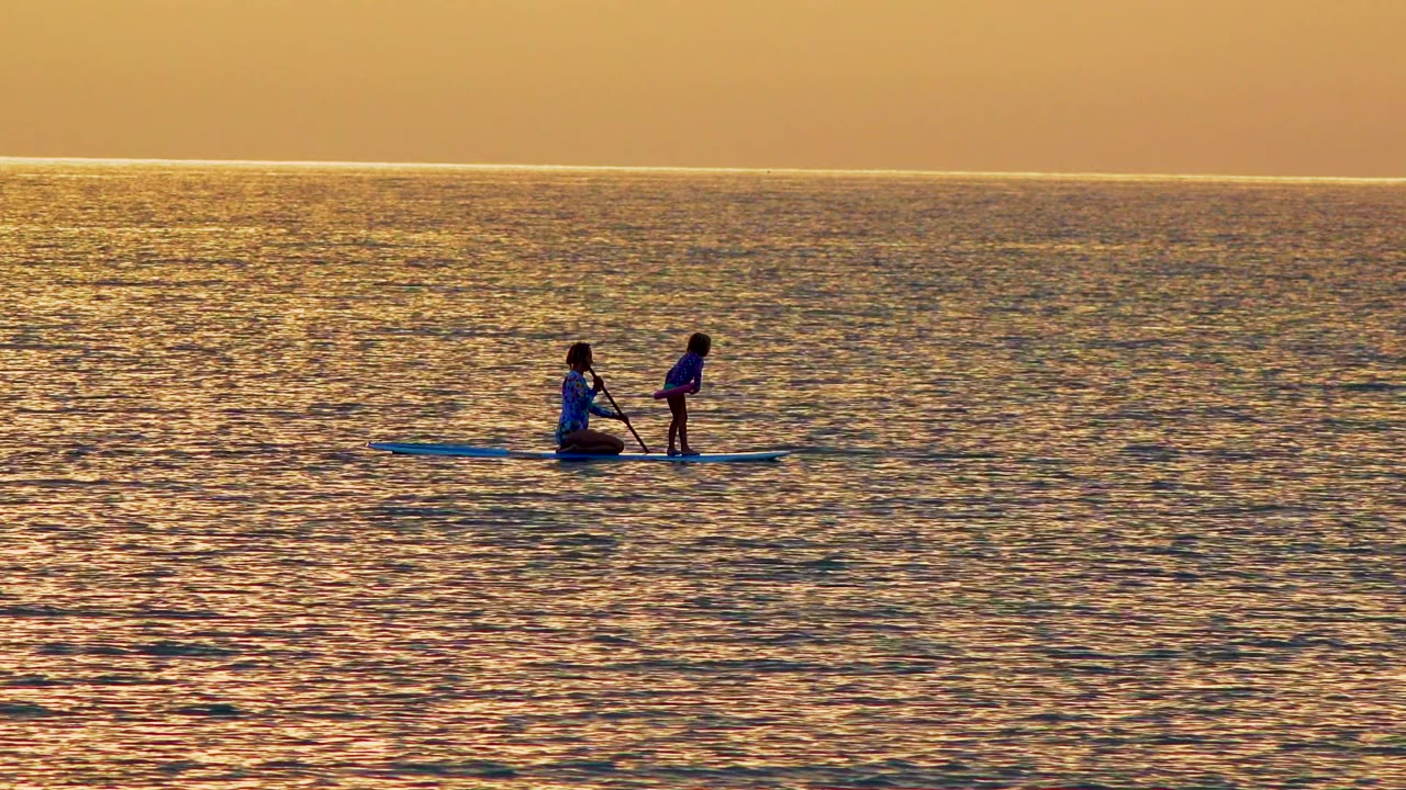 Two silhouetted people ride a paddleboard on sunset water in El Sargento La Ventana, Mexico, with the rear person kneeling and the front person standing upright.
