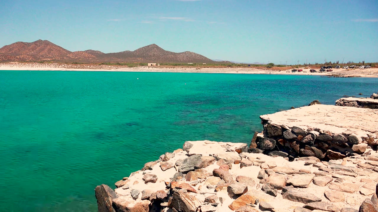 Turquoise coastal water beside pale shoreline rocks and distant mountains.
