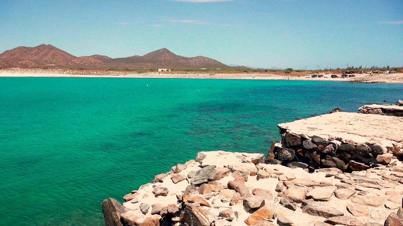 At 0:01, the El Sargento La Ventana shoreline remains framed with bright turquoise water, a rock ledge at right, sandy beach, low mountains, and no visible people.
