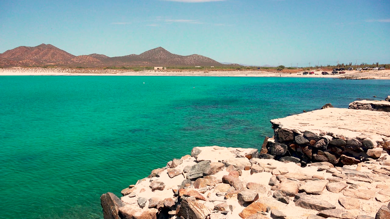 At 0:02, turquoise coastal water fills the center of the El Sargento La Ventana frame, with pale rocks at lower right, beach and mountains behind, and no visible people.