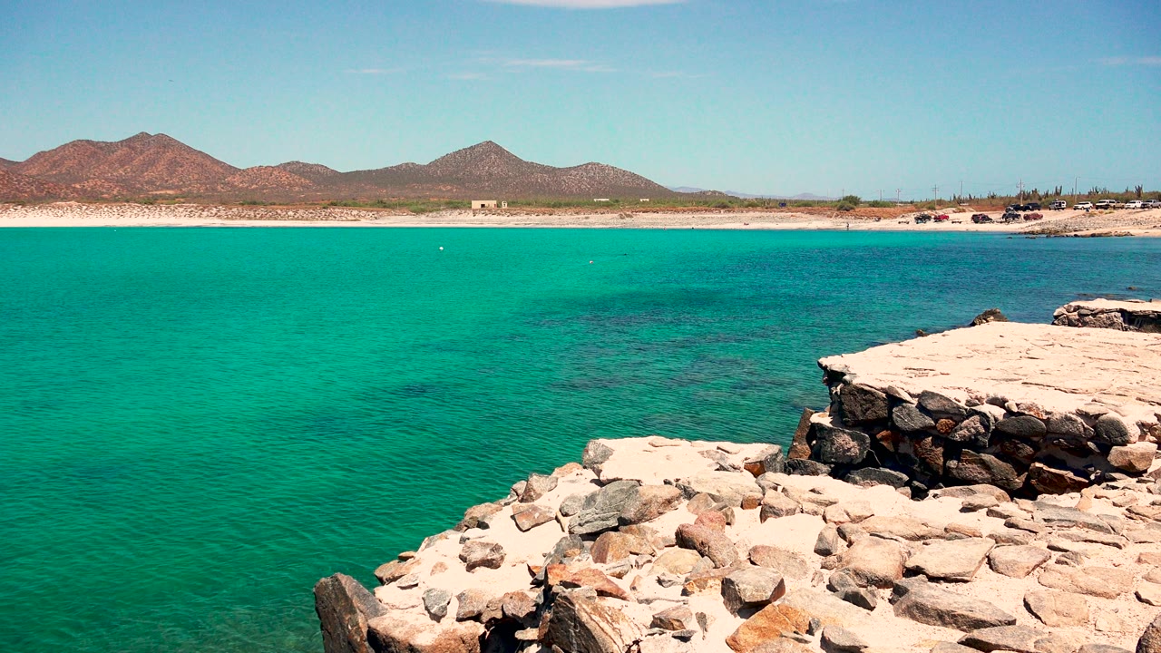 At 0:03, the view opens across turquoise water in El Sargento La Ventana, with foreground stones, a sandy shoreline, distant mountains, and no visible people.