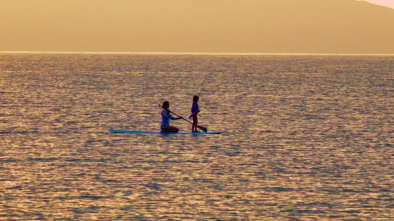 Two people share a blue paddleboard in El Sargento La Ventana, Mexico, with one kneeling near the paddle and the other standing against golden ocean water.