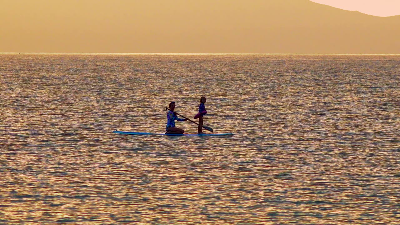 Two silhouetted figures stay on the blue paddleboard in El Sargento La Ventana, Mexico, with the standing person upright and distant mountains layered above the bright horizon.