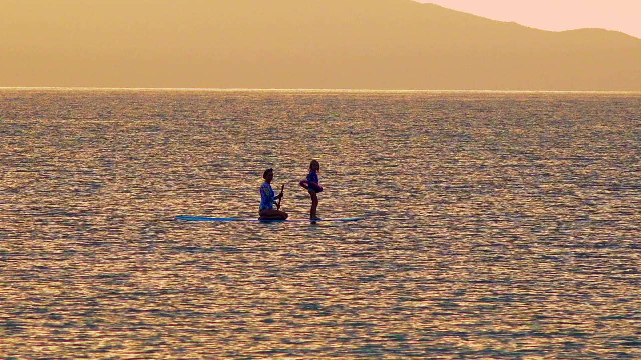 Two people balance on a paddleboard over rippled sunset water.