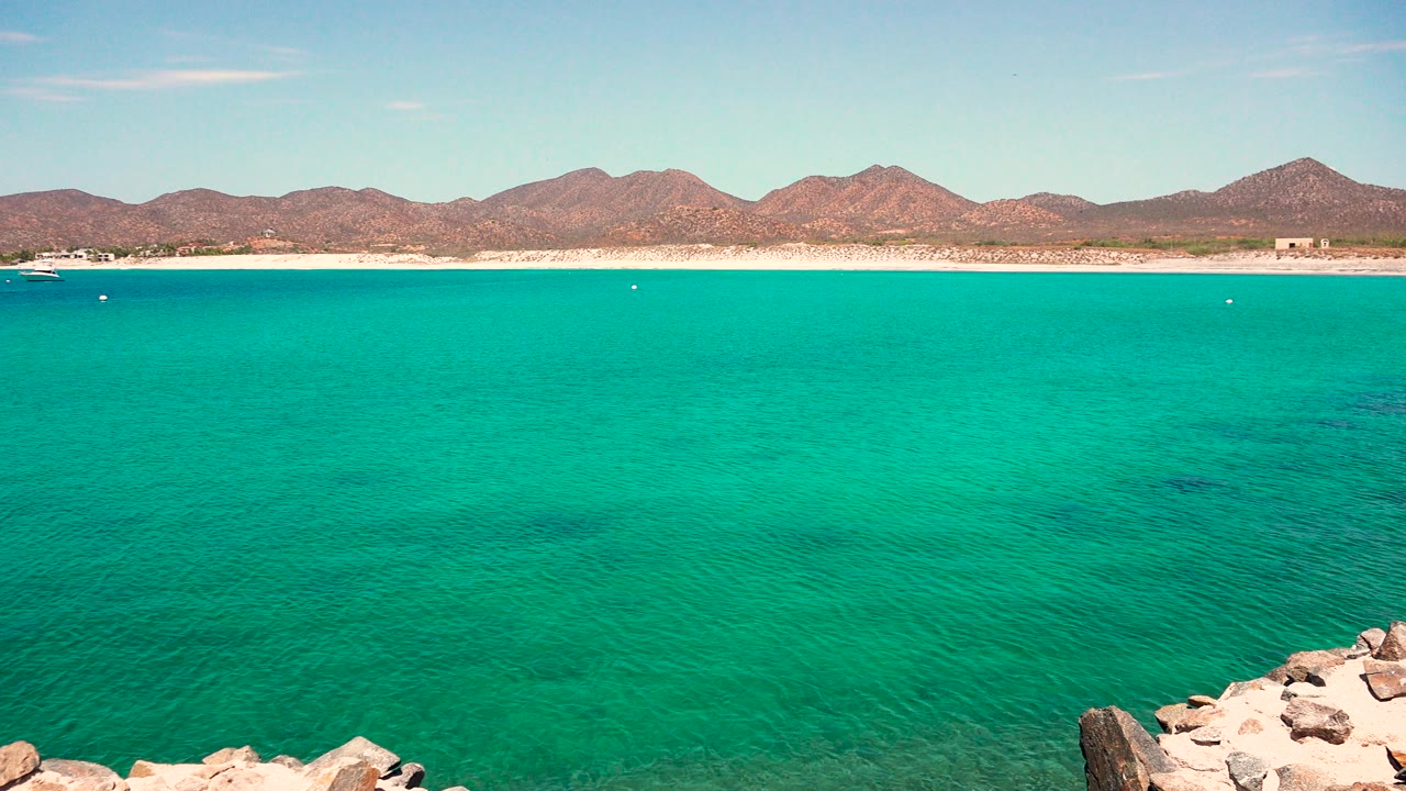Opening frame of turquoise water at El Sargento La Ventana Mexico, with pale foreground rocks, a distant sandy shoreline, brown mountains, blue sky, and no visible people.