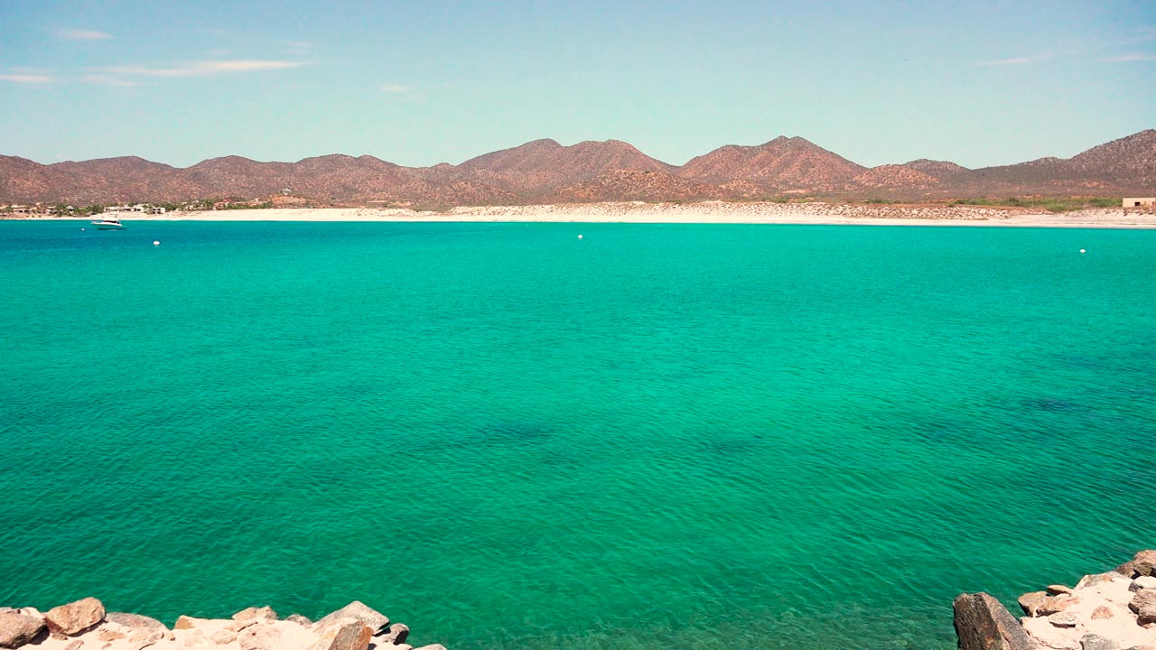 Second frame from El Sargento La Ventana Mexico showing open turquoise water, small distant boats, a pale beach strip, mountain ridgeline, blue sky, and no visible people.