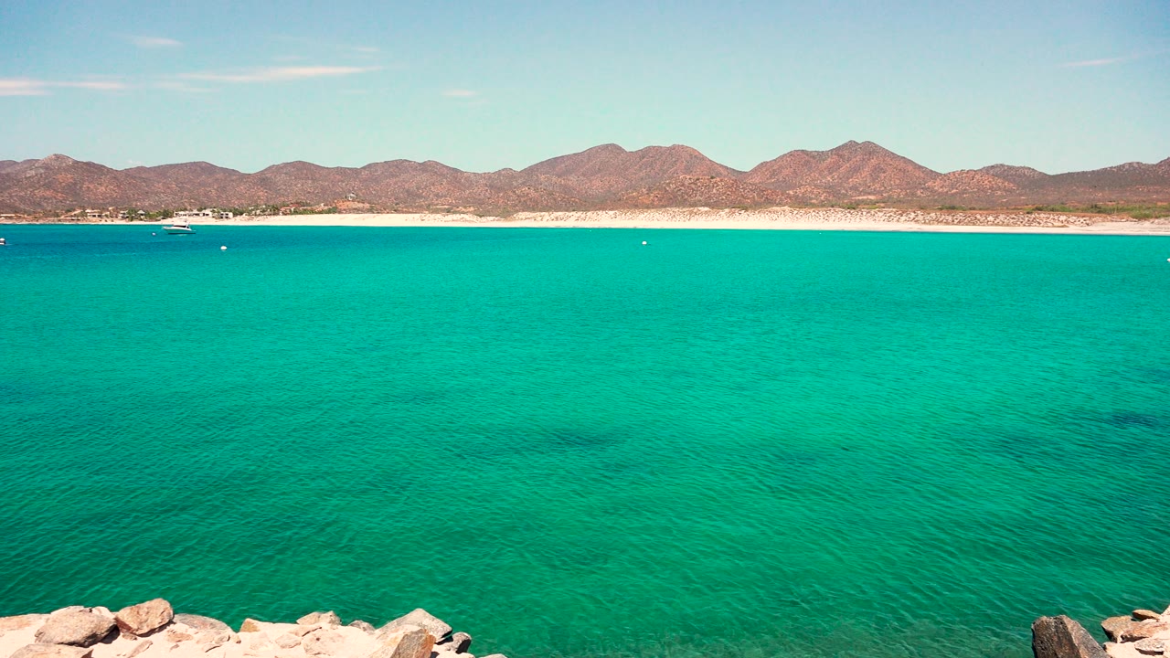 Third frame of the El Sargento La Ventana Mexico coastline, with rocks along the bottom edge, clear turquoise water, a distant beach, mountains, and no visible people.