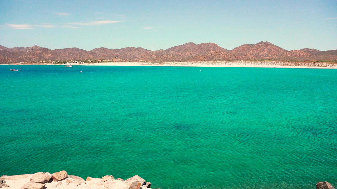Calm turquoise bay with a rocky foreground corner and mountains across the shoreline.