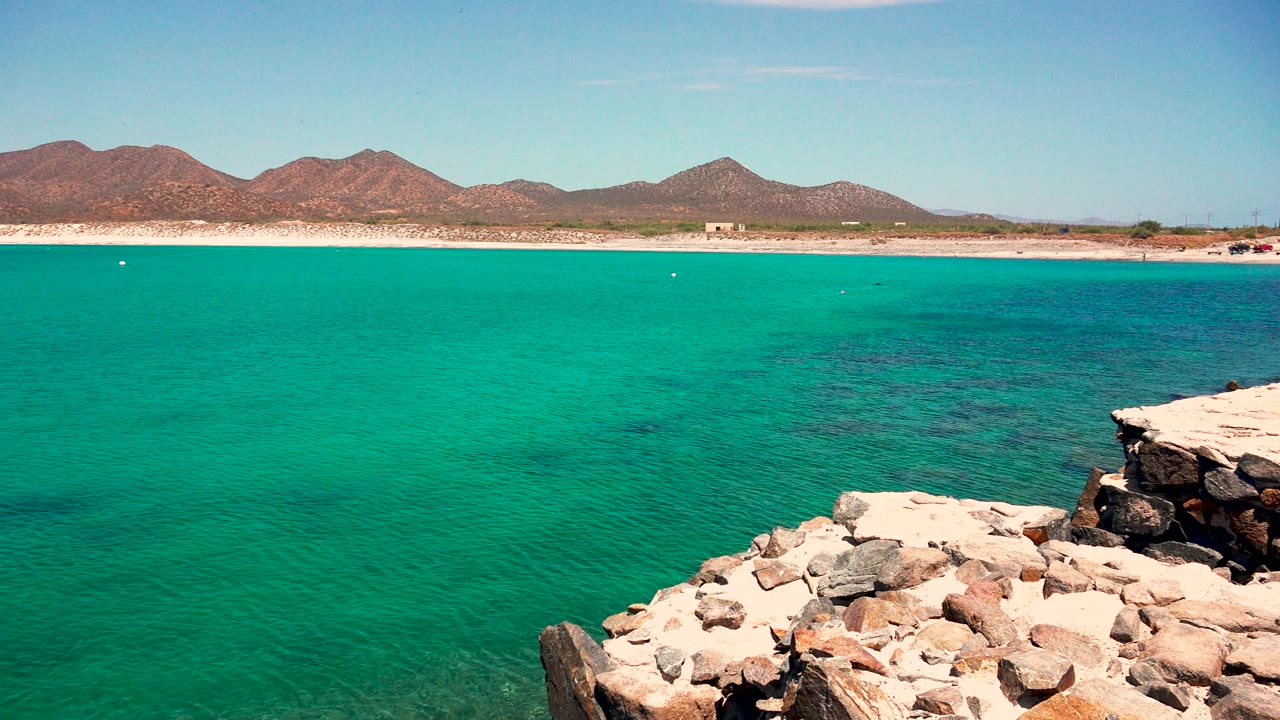 Opening frame of El Sargento La Ventana, Mexico with turquoise ocean, pale rocks at the lower right, a sandy shoreline, distant brown mountains, clear sky, and no visible people.