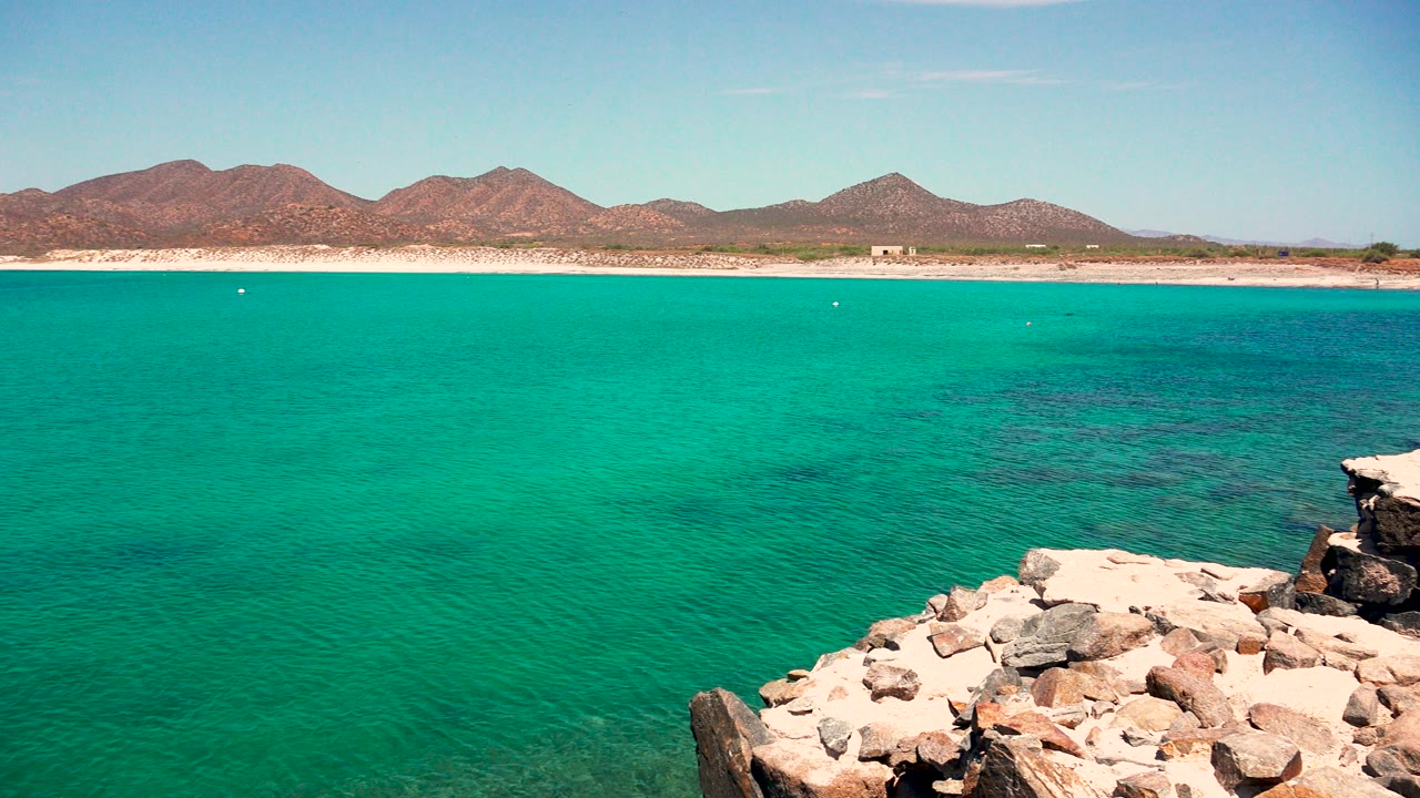 El Sargento La Ventana, Mexico coastline at 0:01, showing more open turquoise water across the center, foreground rocks at the lower right, distant mountains, clear sky, and no visible people.