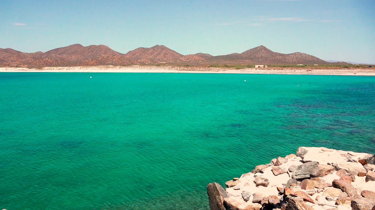 At 0:02 in El Sargento La Ventana, Mexico, the rocky shore sits lower right beside wide turquoise water, with dark underwater patches, a pale beach line, distant mountains, and no visible people.
