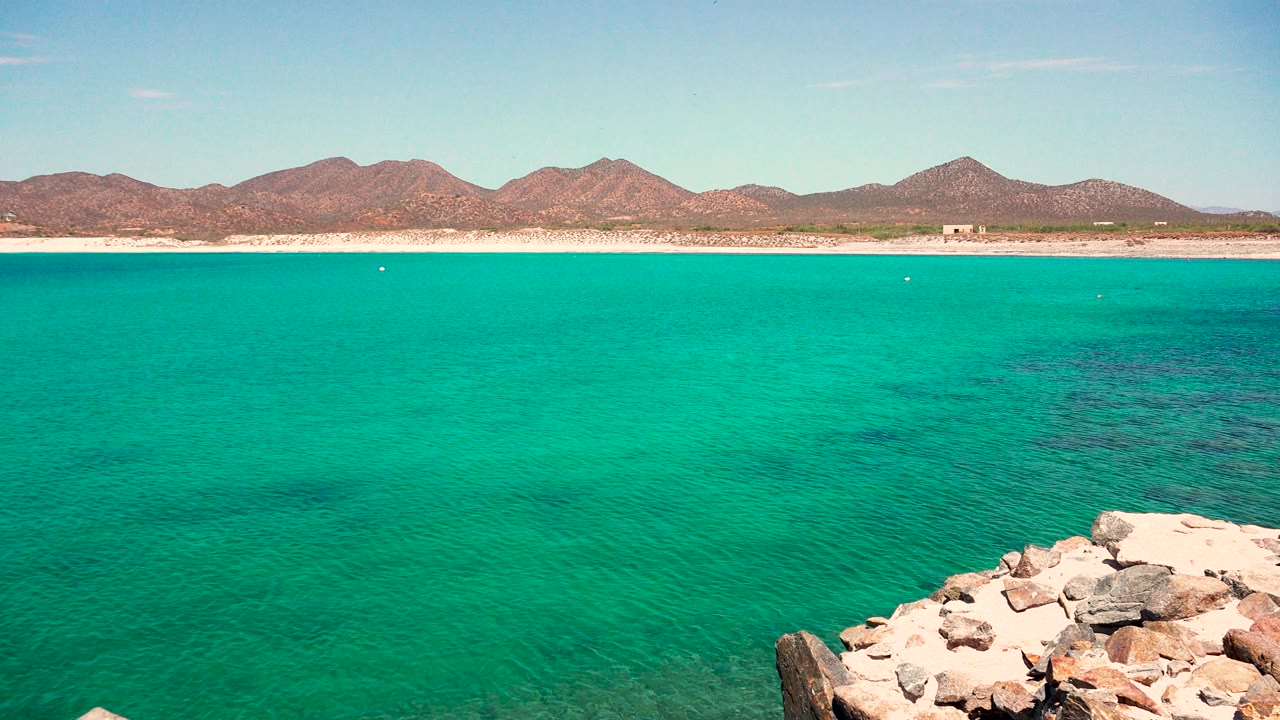 Mid-clip view in El Sargento La Ventana, Mexico with the foreground rocks lower in frame, a broad turquoise water surface, sandy shoreline, low mountains, clear blue sky, and no visible people.