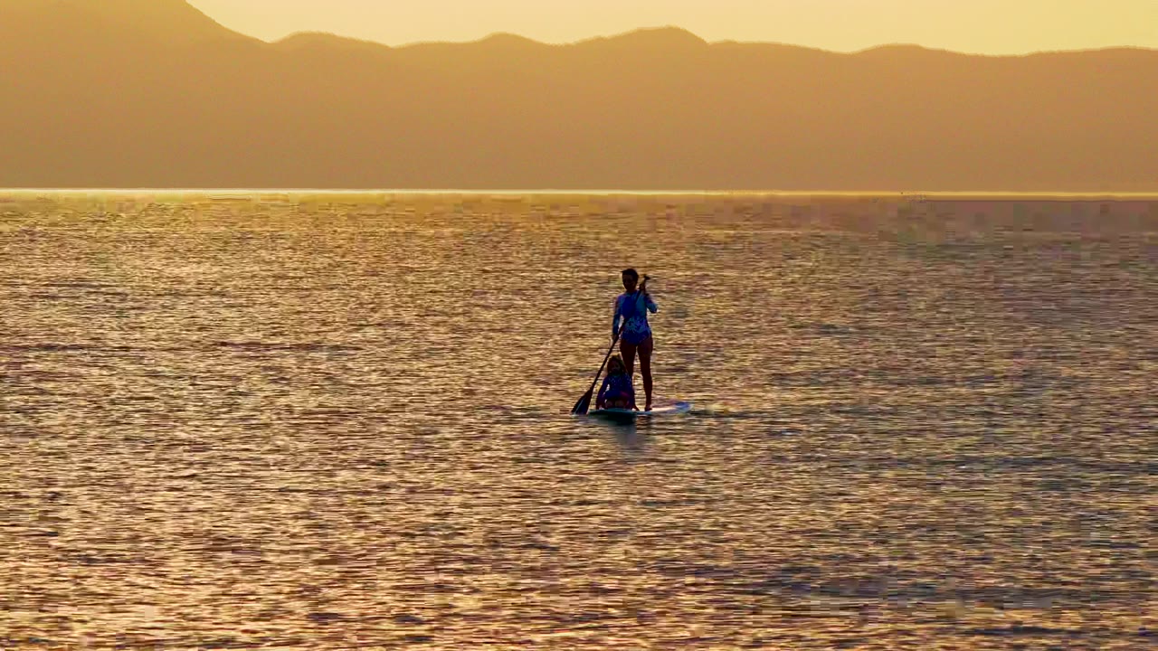 A person stands on a paddleboard on calm golden water in El Sargento La Ventana, Mexico, with a low mountain ridge across the horizon.