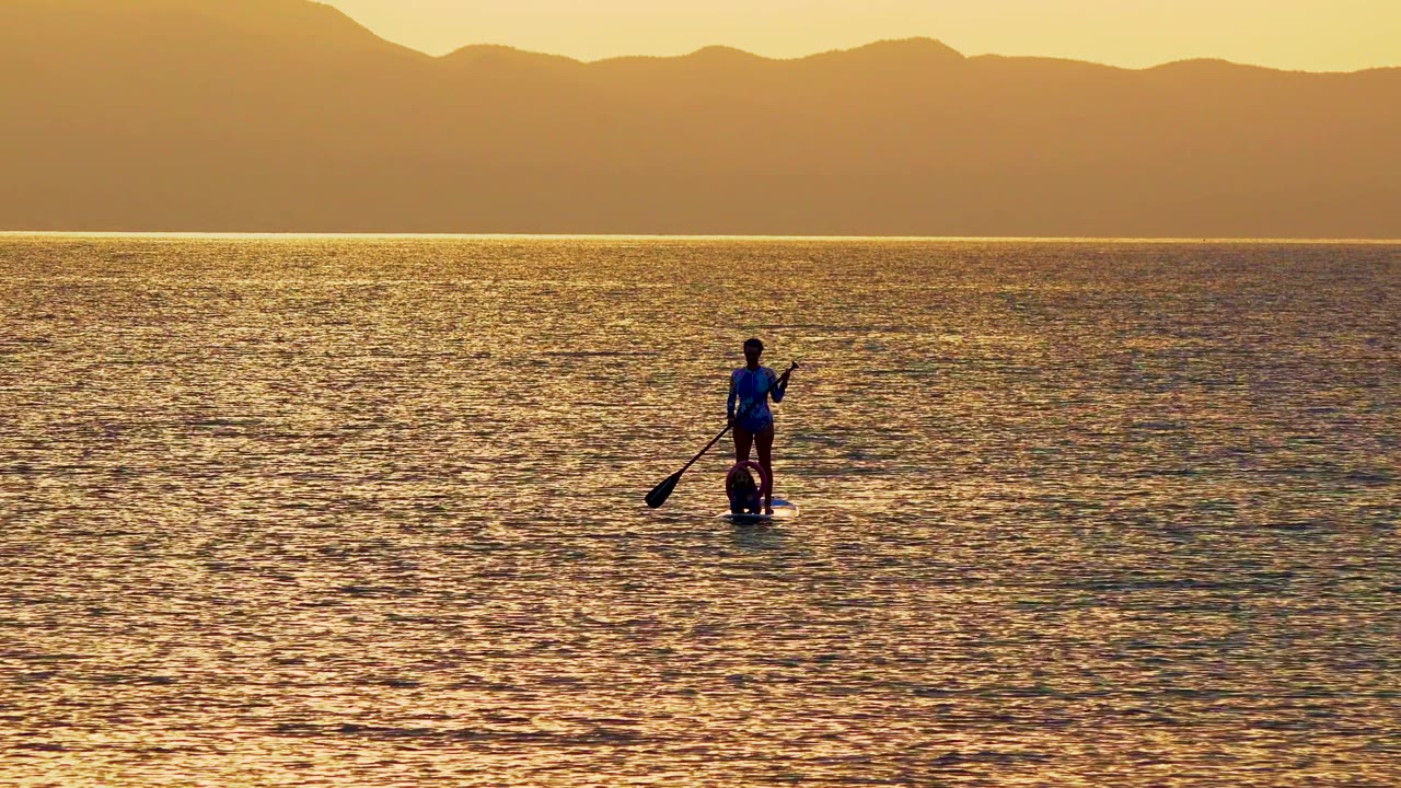 A person stands on a paddleboard in La Ventana, Mexico, with a paddle angled into golden ocean water and distant mountains behind the horizon.