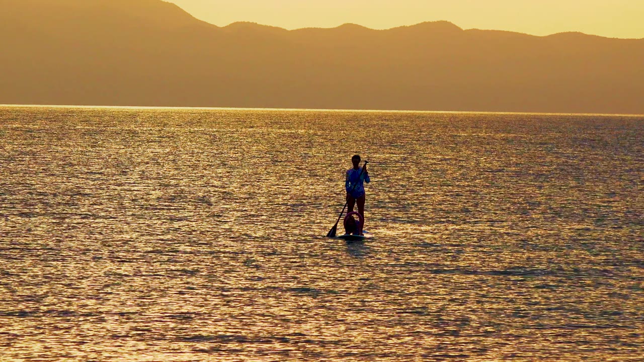 Paddleboarder centered on rippled sunset water beneath distant mountains.