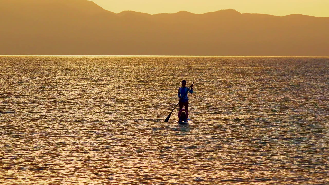 A lone paddleboarder appears toward the right side of the La Ventana frame, silhouetted against shimmering gold water with soft mountains in the background.