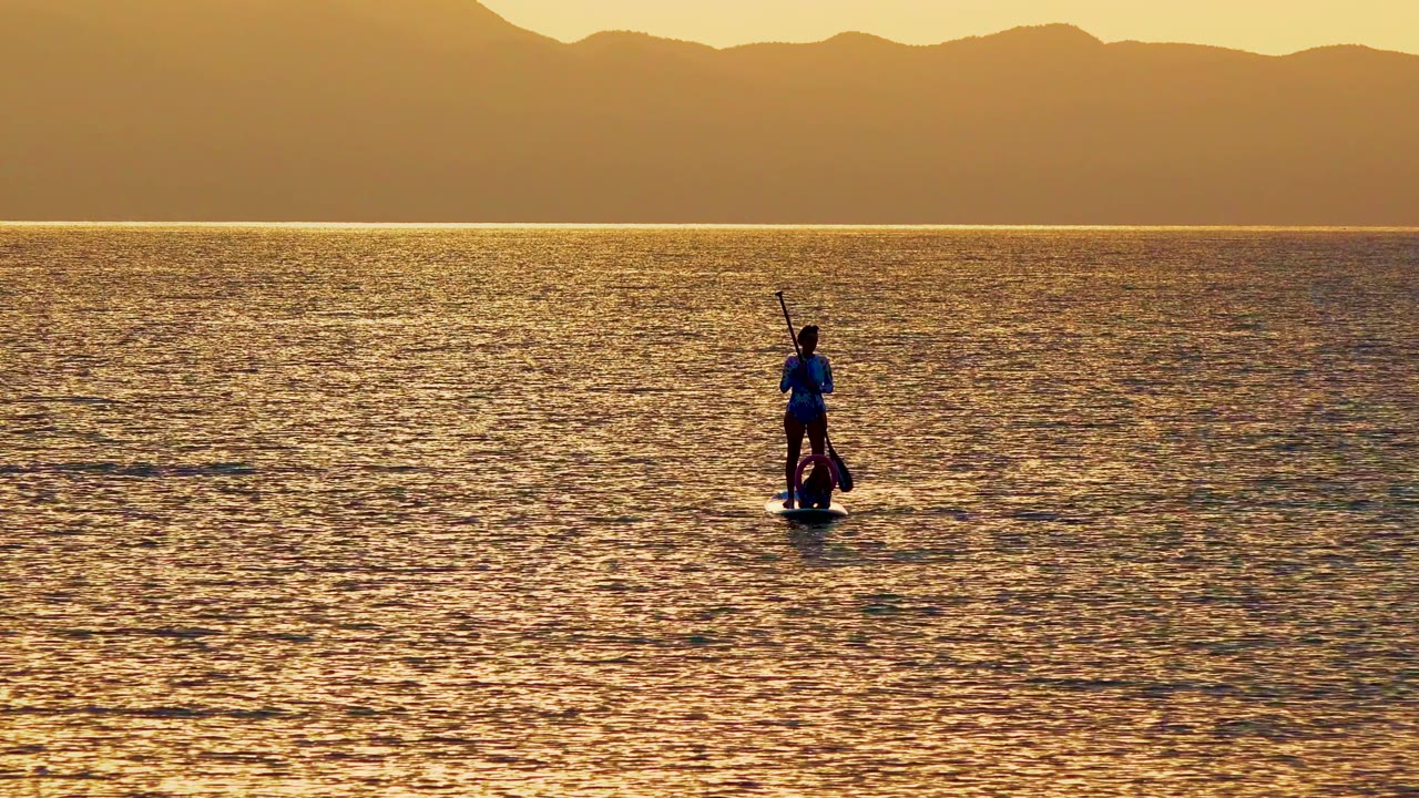 A distant person stands on a paddleboard in La Ventana, Mexico, holding a paddle upright on golden rippled water with mountains along the horizon.