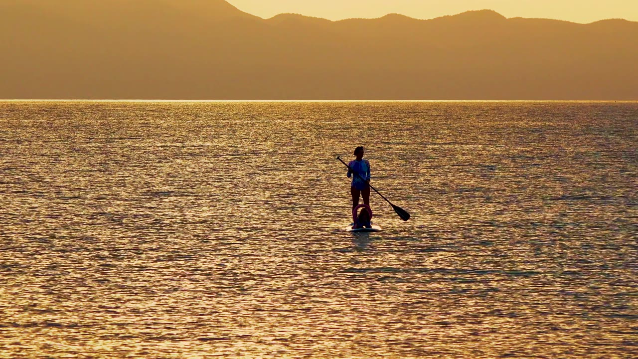 A distant paddleboarder in La Ventana, Mexico, appears on the right side of the wide frame, with the paddle angled outward over golden water and mountains in the background.