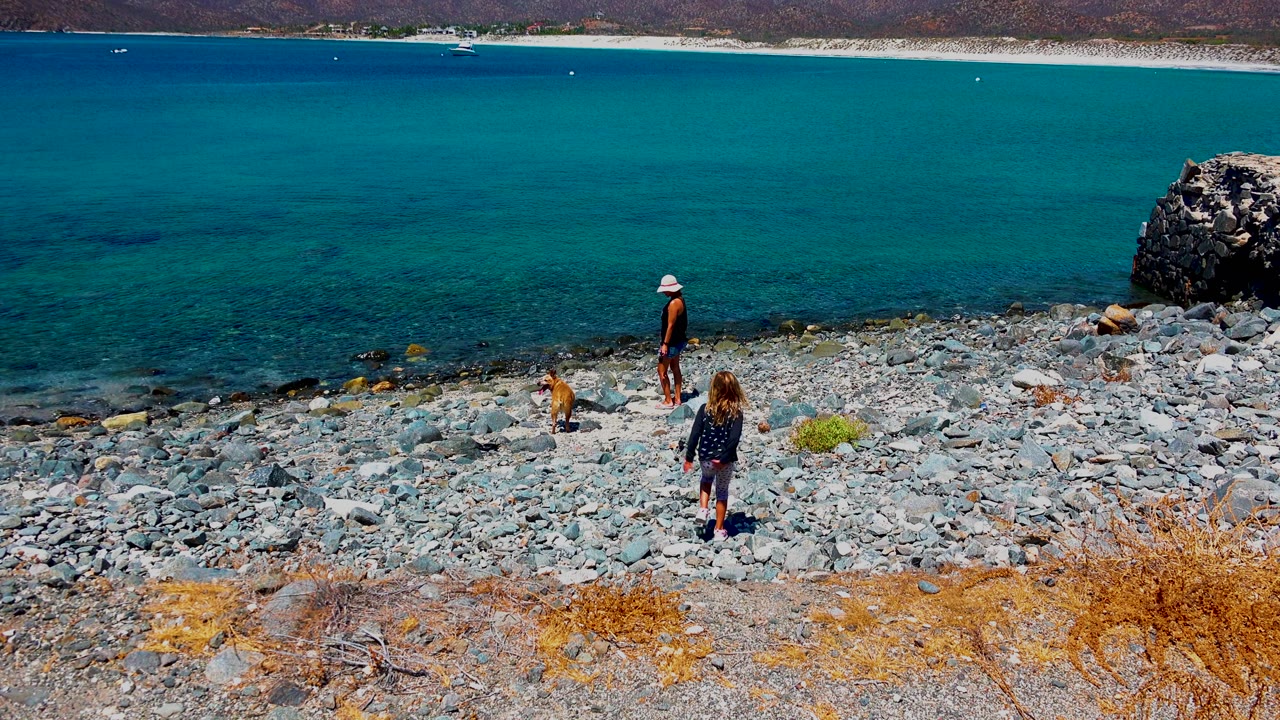At El Sargento, Mexico, the girl is farther down the rocky shore as the adult stands near the tan dog, with blue water filling the background.