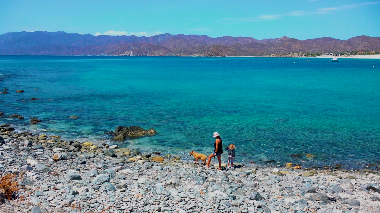 Adult in a white hat, small child, and tan dog walk along the rocky shore in El Sargento, Mexico, with turquoise water and distant mountains behind them.