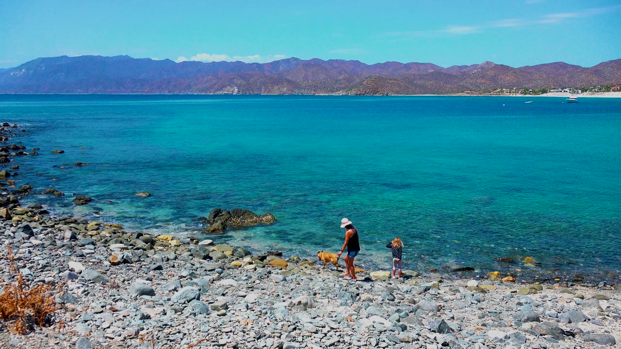 Adult and child near a dog on a rocky shoreline with clear blue water.