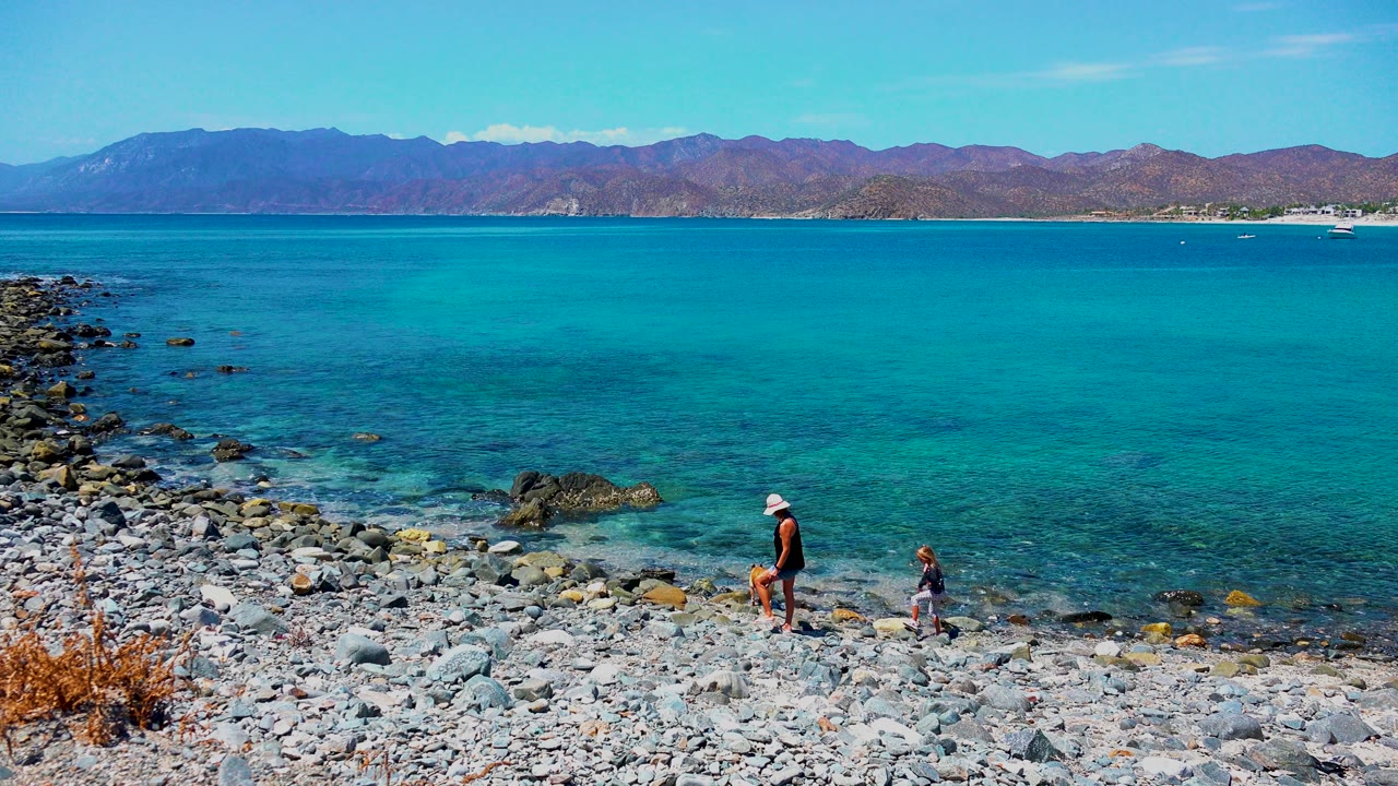 The adult and child are slightly farther right on the rocky El Sargento shore as the tan dog moves near them, with turquoise water filling most of the frame.