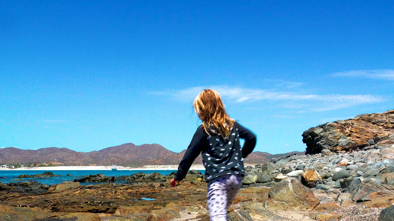 Young girl seen from behind walking onto rocks at the El Sargento shoreline in Mexico, with blue ocean, distant mountains, and a large rock outcrop ahead.