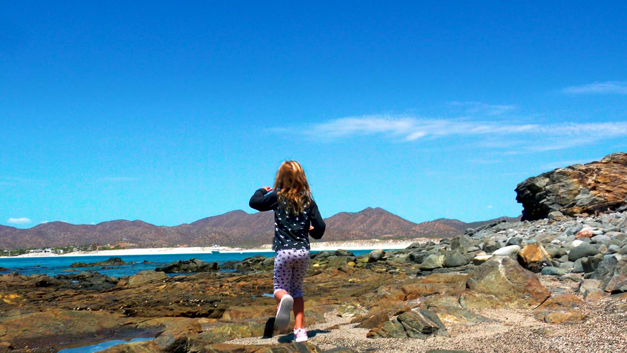 Girl walking farther across rocks toward ocean and mountains.
