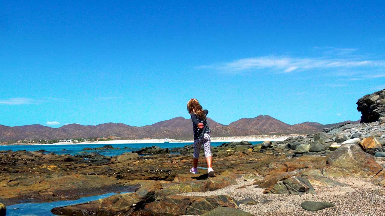 The child appears smaller in the frame on the El Sargento rocky shore, standing near dark tidal rocks with turquoise water, pale sand, and mountain ridges in the background.