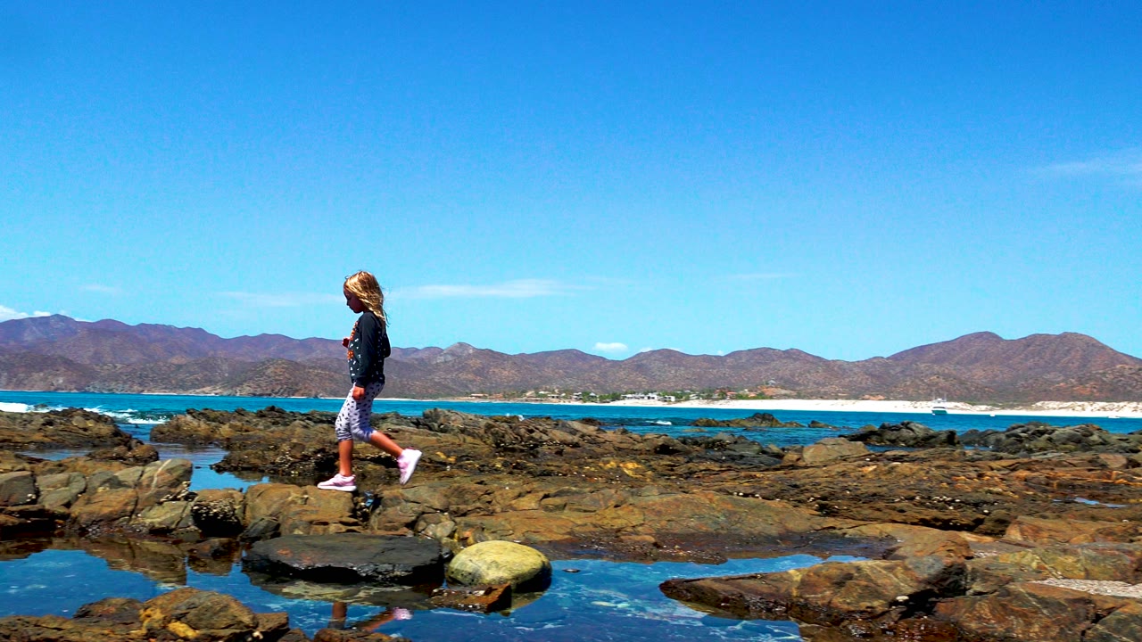 Girl walking on dark rocks beside tidal pools in El Sargento, Mexico, with turquoise ocean and brown mountains under a clear blue sky.
