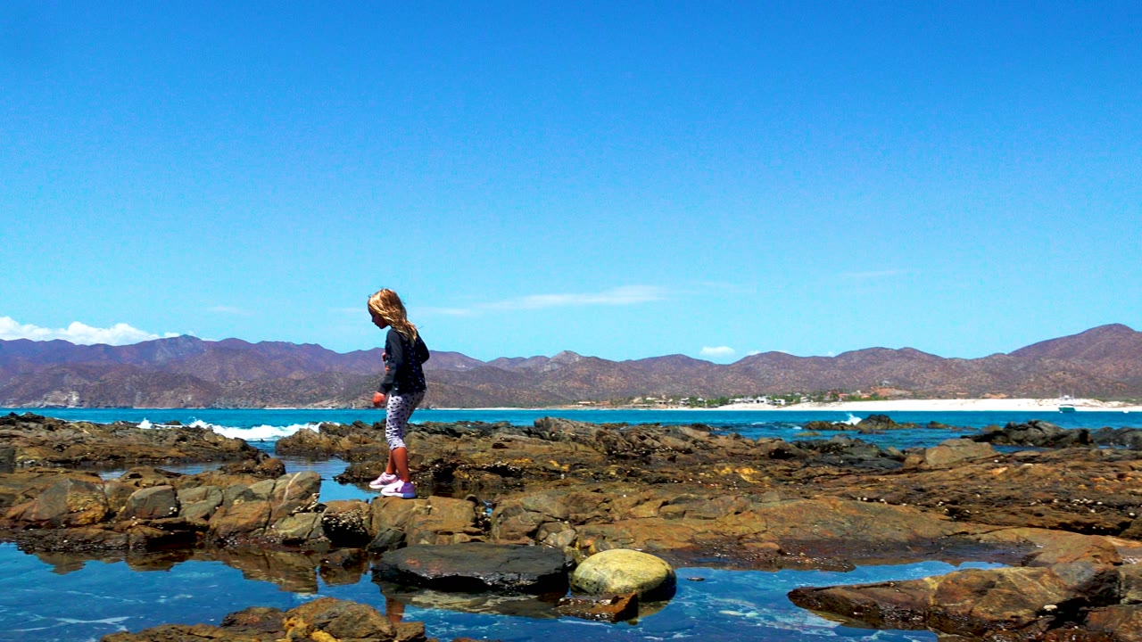 Girl stepping across uneven coastal rocks in El Sargento, Mexico, with reflective tide pools, low surf, distant mountains, and open sky behind her.