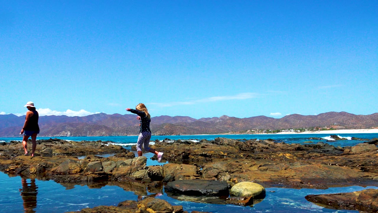 Man in a hat stands on the rocky shoreline in El Sargento, Mexico, facing the water with tide pools, ocean, mountains, and clear sky visible.